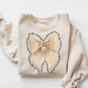 May include: A beige sweatshirt with a brown bow design. The bow is surrounded by the words "Beautiful, Grateful, Blessed" repeated in a circle.