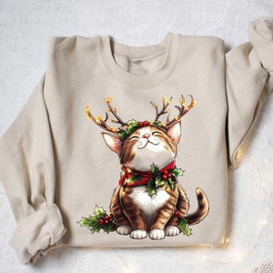 May include: A beige sweatshirt with a Christmas-themed graphic of a cat wearing a reindeer antler headband with lights and a red scarf with holly.