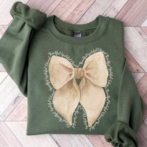 May include: A green sweatshirt with a large brown bow on the front. The bow is surrounded by the words "Thankful Grateful Blessed" repeated around the bow.