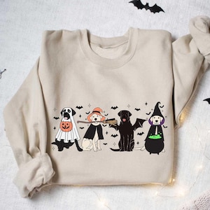 May include: Beige sweatshirt featuring a Halloween-themed graphic of four dogs in costumes: a ghost, a witch, a bat, and a witch with a cauldron. Black bats and stars are scattered around the dogs. A cozy, festive design.