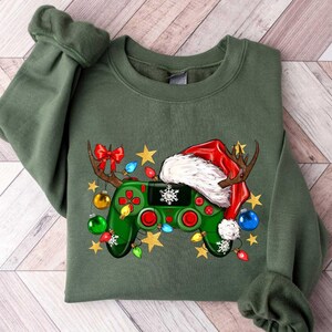 May include: Green sweatshirt with a festive design featuring a video game controller decorated with a Santa hat, reindeer antlers, and Christmas lights. The controller has red buttons and a snowflake. Gold stars are scattered around the design.