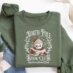 May include: Green sweatshirt with a white graphic design that says "North Pole Book Club" with a mug of hot cocoa and a gingerbread man. The text below the graphic says "Hot Cocoa Served Daily".