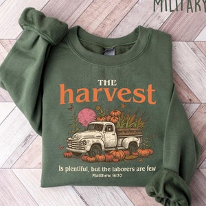 May include: A green sweatshirt with the text "THE harvest" in orange letters. The sweatshirt features a vintage truck filled with pumpkins and fall foliage. The text "Is plentiful, but the laborers are few Matthew 9:37" is printed below the truck.