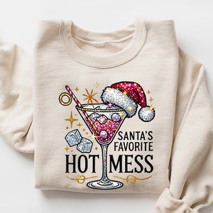 May include: A beige sweatshirt with a festive design featuring a martini glass with a red liquid, ice cubes, and a red Santa hat. The text "Santa's Favorite Hot Mess" is printed in gold glitter.