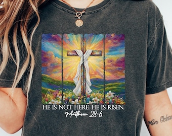 May include: A charcoal grey t-shirt with a colorful painting of a cross with a white cloth on it. The painting is set against a sunset background with a field of flowers in the foreground. The text "HE IS NOT HERE HE IS RISEN. Matthew 28:6" is printed below the painting.