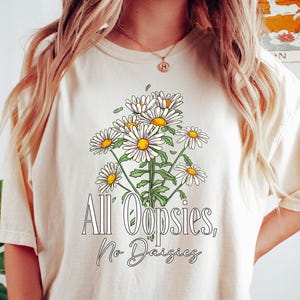 May include: Cream-colored t-shirt featuring a daisy bouquet graphic with the text "All Oopsies, No Daisies" in a script font. The daisies have white petals and yellow centers. The shirt is a casual, comfortable style.