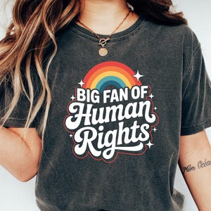 May include: Dark gray t-shirt with the text "BIG FAN OF HUMAN RIGHTS" in a retro font, above a rainbow graphic. The shirt is worn with blue jeans and white sneakers. The overall theme is social justice.