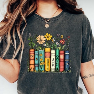 Peut inclure: Un t-shirt gris fonc&eacute; avec un motif repr&eacute;sentant une rang&eacute;e de livres color&eacute;s avec les mots "Tango", "Delta", "Fox Trot" et "8647" visibles. Les livres sont entour&eacute;s de feuilles vertes et de fleurs jaunes, orange et rouges.