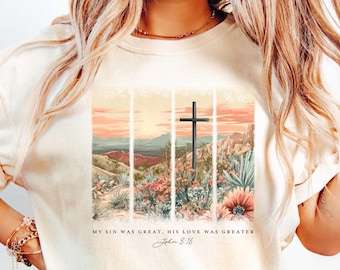 Puede incluir: Una camiseta beige con un gráfico de un paisaje de puesta de sol y una cruz. El texto "My sin was great, His love was greater John 3:16" está impreso en la camiseta.