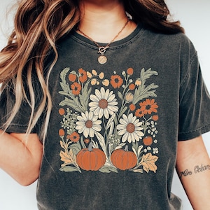 May include: A dark grey t-shirt with a vintage floral print featuring white daisies, orange pumpkins, and green leaves. The shirt has a crew neck and short sleeves.
