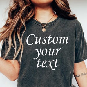 May include: Dark gray t-shirt with the text "Custom your text" in white. The shirt is worn with blue jeans and white sneakers. The image shows a casual, relaxed style.