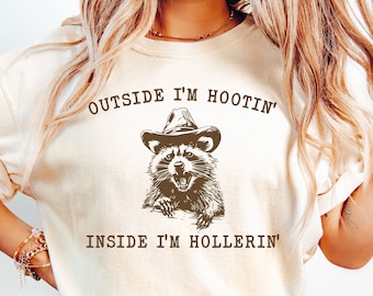 Op de afbeelding: Een wit T-shirt met een bruine wasbeer die een cowboyhoed draagt en de tekst "Outside I'm Hootin' Inside I'm Hollerin'"