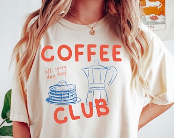 May include: Cream-colored t-shirt with the words "COFFEE CLUB" in red, along with a blue illustration of a coffee pot and pancakes. The phrase "all every day day" is printed above the pancakes. The shirt is a casual, comfortable style.