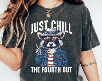 May include: Dark gray t-shirt with the text "JUST CHILL THE FOURTH OUT". The graphic features a raccoon wearing heart-shaped sunglasses, a cowboy hat, and a denim jacket. The raccoon is giving a peace sign.