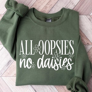 May include: An olive green sweatshirt with the text "ALL OOPSIES no daisies" in white script. The text is decorated with small daisy flower graphics. The sweatshirt is folded neatly on a light wood-grain surface.
