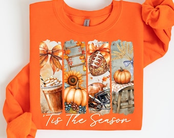May include: Orange sweatshirt with a fall-themed graphic. The graphic includes a bow, a latte, a sunflower, a pumpkin, a football, and the text "Tis The Season". The sweatshirt is made of a soft material.