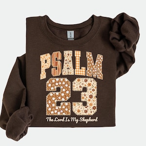 May include: A brown sweatshirt with the text "PSALM 23" in a floral and plaid design. The number "23" is also in a floral pattern. The phrase "The Lord Is My Shepherd" is printed below the numbers.