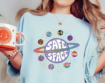 Può includere: Una maglietta blu chiaro con una grafica di un pianeta viola con il testo "SAFE SPACE" in lettere arcobaleno. Il pianeta è circondato da un anello con pianeti più piccoli in vari colori e motivi.
