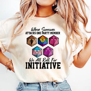 May include: Cream-colored t-shirt with the text "When Someone Attacks One Party Member, We All Roll For Initiative." The shirt features six colorful dice with various symbols. The person is wearing blue jeans.