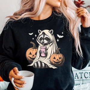 Puede incluir: Sudadera negra con un gr&aacute;fico de un mapache vestido de fantasma y sosteniendo una bebida rosa. El mapache est&aacute; rodeado de calabazas.