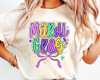 May include: A light beige t-shirt with a purple, green and yellow graphic that says "Mardi Gras" with a purple bow and glitter accents.