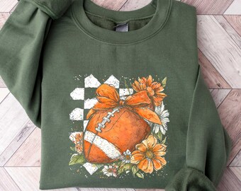 May include: Olive green sweatshirt featuring a football design with a bow and floral accents. The football is orange with white stripes, set against a checkered background. The flowers are orange and white. The sleeves are cuffed.