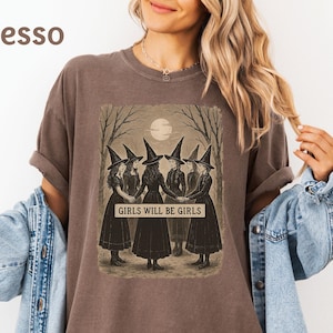 K&ouml;nnte beinhalten: Espressofarbenes T-Shirt mit einer Illustration im Vintage-Stil von f&uuml;nf Hexen in schwarzen Kleidern und spitzen H&uuml;ten, die sich unter einem Vollmond an den H&auml;nden halten. Der Text "GIRLS WILL BE GIRLS" ist darunter gedruckt.