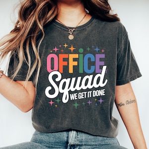 May include: Dark gray t-shirt with the words "OFFICE Squad WE GET IT DONE" in colorful, bold lettering. The shirt features a crew neck and short sleeves. The person is wearing blue jeans and white sneakers.