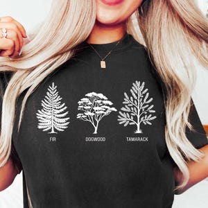 Pu&ograve; includere: Maglietta nera con una grafica bianca di tre alberi. Gli alberi sono etichettati "Fir", "Dogwood" e "Tamarack".