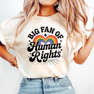 May include: Cream-colored t-shirt with the text "BIG FAN OF Human Rights" in black script and a rainbow heart design. The shirt is paired with light blue denim jeans.