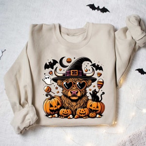May include: A beige sweatshirt with a Halloween-themed design featuring a Highland cow wearing a witch's hat and sunglasses, surrounded by pumpkins, bats, and candy.