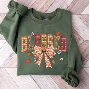 May include: An olive green sweatshirt with the word "BLESSED" in a patchwork design. The letters feature plaid, leopard print, and floral patterns. A large bow and fall leaves and flowers are below the word.