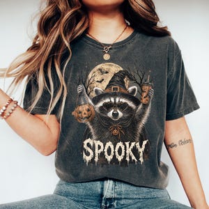 May include: A charcoal gray t-shirt featuring a graphic of a raccoon wearing a witch hat and sunglasses, holding a pumpkin bucket and a drink. The raccoon is set against a full moon and bare trees, with the word SPOOKY printed in a distressed font.