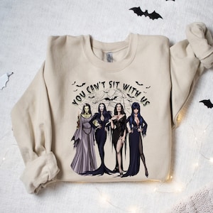 May include: A beige sweatshirt with a graphic print of four women dressed in black and purple gowns. The text "You can't sit with us" is printed in a green circle around the women. The background of the graphic is a black spiderweb with bats flying around it.