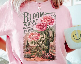 May include: Pink t-shirt with a vintage-style graphic of roses and a cactus. The graphic says "Bloom Where You Are Planted".