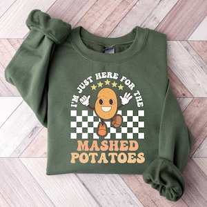May include: A dark green sweatshirt with the text "I'M JUST HERE FOR THE MASHED POTATOES" and a cartoon potato character. The design includes a checkered pattern and stars. The sweatshirt is laid flat on a wooden surface.