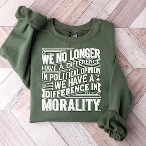 May include: An olive green sweatshirt with the text "WE NO LONGER HAVE A DIFFERENCE IN POLITICAL OPINION WE HAVE A DIFFERENCE IN MORALITY" in white lettering. The sweatshirt is folded on a light wood surface.