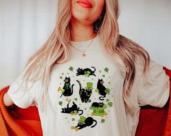 Puede incluir: Una camiseta blanca con un diseño de gato negro. Los gatos llevan sombreros verdes y sostienen tréboles. El diseño está rodeado de tréboles verdes y monedas de oro.