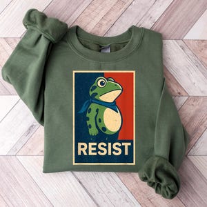 May include: A green sweatshirt featuring a frog illustration with the word "RESIST" in a vintage-style poster design. The frog is green with a blue scarf. The background of the poster is red and blue.