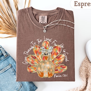 May include: Espresso-colored t-shirt with a turkey graphic. The turkey has a colorful feather design and a bow. The text "Give Thanks To The Lord For He Is Good" and "Psalm 136:1" are printed on the shirt. A silver necklace is draped on the shirt.