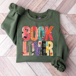 May include: An olive green sweatshirt with the words "BOOK LOVER" in a colorful patchwork design. The letters are made up of various patterns and colors, including red, blue, and yellow. The sweatshirt has long sleeves and a crew neck.