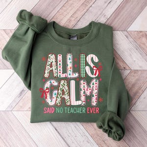 May include: Green crewneck sweatshirt with the text "ALL IS CALM SAID NO TEACHER EVER." The text is in a variety of festive patterns and colors, including red, white, and green. The sweatshirt has long sleeves and a ribbed collar and cuffs.