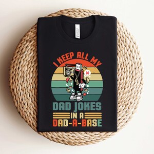 May include: Black t-shirt with a colorful graphic. The graphic features a cartoon USB drive character with a boombox, coins, and the text "I KEEP ALL MY DAD JOKES IN A DAD-A-BASE."