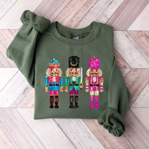 May include: Green sweatshirt with a festive design of three nutcracker soldiers. The nutcrackers are wearing colorful costumes with gold accents and glitter details.