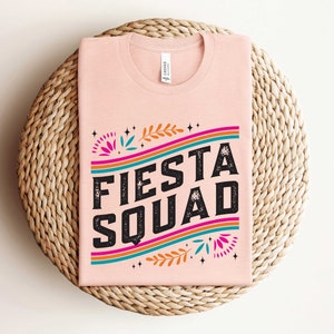May include: A pink t-shirt with the text "Fiesta Squad" in black letters with a colorful design of flowers and stripes.
