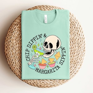 Pu&ograve; includere: Una maglietta verde chiaro con una grafica di uno scheletro che beve una margarita e il testo "Chip Dippin' & Sippin' Margarita".