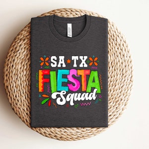 May include: A dark gray t-shirt with a colorful graphic design that says "SA TX Fiesta Squad". The graphic is in a festive style with colorful letters and decorative elements.