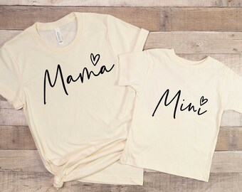 May include: Two cream-colored t-shirts with black script. The larger shirt says "Mama" with a small heart. The smaller shirt says "Mini" with a small heart.