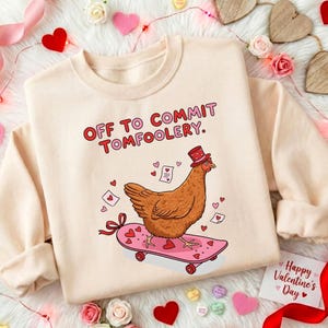K&ouml;nnte beinhalten: Ein cremefarbenes Sweatshirt mit dem Text "OFF TO COMMIT TOMFOOLERY." Ein Cartoon-Huhn mit Hut f&auml;hrt auf einem rosa Skateboard mit Herzen. Das Design umfasst Herzen und Valentinstagselemente.