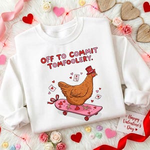May include: White sweatshirt with the text "OFF TO COMMIT TOMFOOLERY." A cartoon chicken wearing a top hat rides a pink skateboard decorated with hearts. The design includes hearts and Valentine's Day elements.